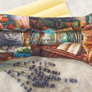 Op de afbeelding: Een lange, rechthoekige lavendel-geurende warmtepack met een kleurrijke print van katten, boeken en een venstersc&egrave;ne. De pack ligt op een wit oppervlak met een bosje lavendeltakjes op de voorgrond.