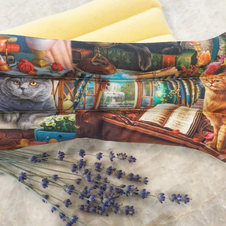 Op de afbeelding: Een lange, rechthoekige lavendel-geurende warmtepack met een kleurrijke print van katten, boeken en een venstersc&egrave;ne. De pack ligt op een wit oppervlak met een bosje lavendeltakjes op de voorgrond.