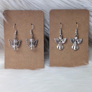 K&ouml;nnte beinhalten: Zwei Paar Ohrringe mit Engelsmotiven. Ein Paar hat klare, facettierte Perlen und detaillierte Fl&uuml;gel. Das andere Paar hat ein einfacheres Design mit einem kleinen Kristallakzent. Beide Sets sind silberfarben und h&auml;ngen an einem einfachen Haken.