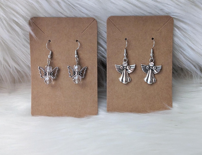 K&ouml;nnte beinhalten: Zwei Paar Ohrringe mit Engelsmotiven. Ein Paar hat klare, facettierte Perlen und detaillierte Fl&uuml;gel. Das andere Paar hat ein einfacheres Design mit einem kleinen Kristallakzent. Beide Sets sind silberfarben und h&auml;ngen an einem einfachen Haken.