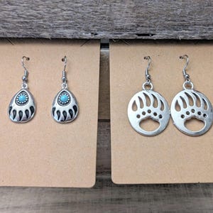 May include: Two pairs of silver bear paw earrings. One pair features a turquoise stone in the center of each paw print. The other pair features a hollow bear paw print.