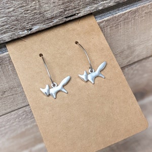 May include: A pair of silver-toned fox-shaped earrings. Each earring features a stylized fox pendant suspended from a curved ear wire. The earrings are displayed on a brown card against a wooden background.