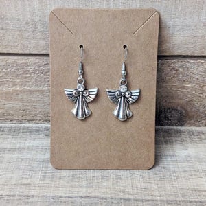 K&ouml;nnte beinhalten: Ein Paar silberfarbene Engels-Ohrringe, pr&auml;sentiert auf einer braunen Karte. Jeder Ohrring zeigt ein Engel-Design mit detaillierten Fl&uuml;geln und einem kleinen, klaren Edelstein. Die Ohrringe h&auml;ngen an silberfarbenen Haken.