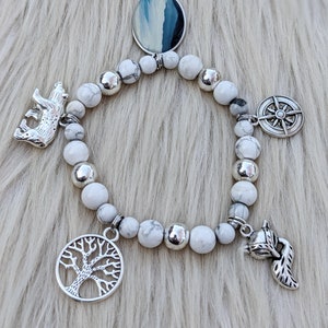 May include: White beaded bracelet with silver charms, including a compass, a tree, a bear, a fox, and a round charm with a blue and white landscape image.