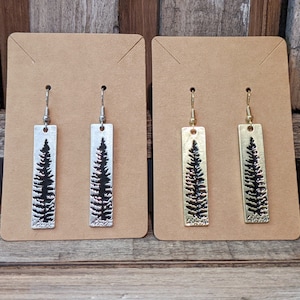 May include: Two pairs of dangle earrings with a rectangular design featuring a silhouette of a pine tree. One pair is silver and the other is gold.