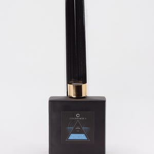 May include: A black reed diffuser with a gold-colored collar and a label that reads "OURCHETMEICA". The diffuser includes black reeds in a clear plastic tube. The label also says "MIDWINTER".