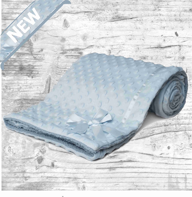 May include: A light blue baby blanket with a white satin ribbon and a bow. The blanket is made of soft, plush fabric and has a textured pattern.