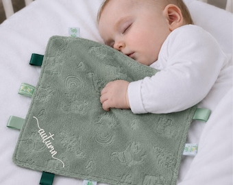 May include: A square sage green baby security blanket featuring an embossed pattern of dinosaurs and rainbows. The fabric is textured with multiple fabric ribbon tags along the edges. The name autumn is embroidered in white cursive script on the front.