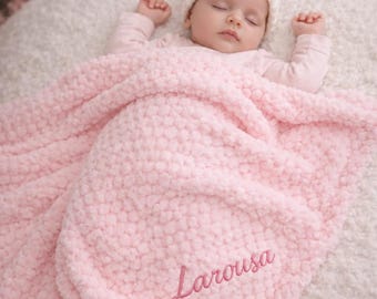 May include: A baby wearing a white knit beanie and a light pink long-sleeved shirt, lying under a soft, textured light pink blanket. The blanket features the name Larousa embroidered in a darker pink script font on the lower right side.