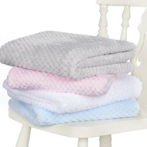 May include: A stack of four soft, textured blankets in pastel colors: gray, pink, white, and light blue. The blankets are folded neatly and stacked on top of each other. The blankets are on a white chair.