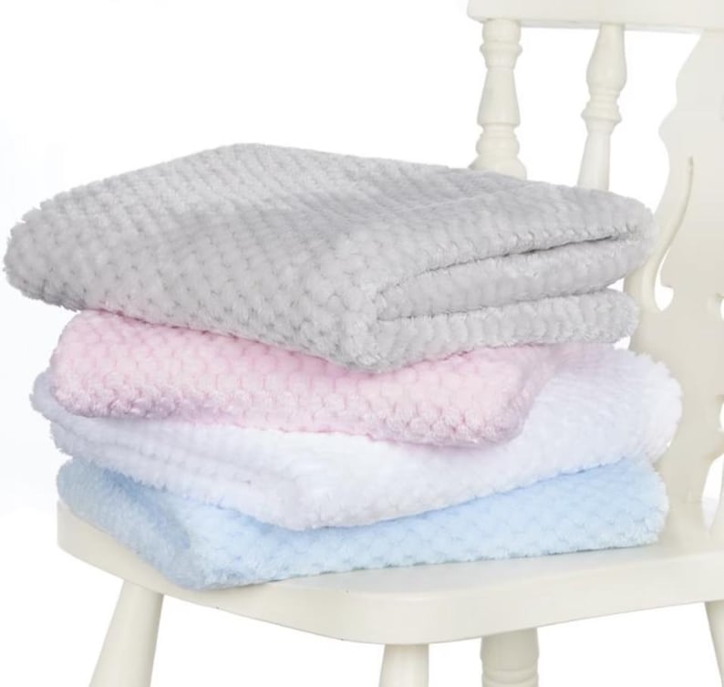 May include: A stack of four soft, textured blankets in pastel colors: gray, pink, white, and light blue. The blankets are folded neatly and stacked on top of each other. The blankets are on a white chair.