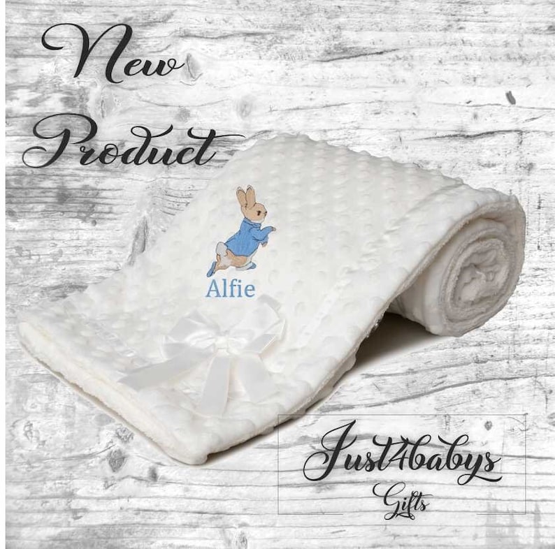 May include: White baby blanket featuring a blue and brown Peter Rabbit design with the name "Alfie" embroidered on it. The blanket is rolled up and tied with a white satin ribbon. The text "Just4babys Gifts" is visible in the bottom right corner.