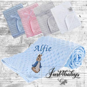 May include: A blue baby blanket with a white embroidered Peter Rabbit design and the name "Alfie". The blanket is made of soft, textured fabric and is perfect for cuddling.