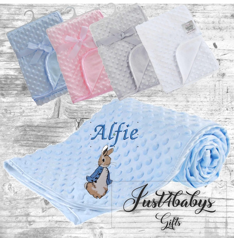 May include: A blue baby blanket with a white embroidered Peter Rabbit design and the name "Alfie". The blanket is made of soft, textured fabric and is perfect for cuddling.