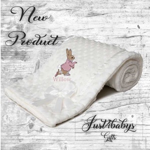 May include: White baby blanket with a white satin bow and a pink embroidered Peter Rabbit with the name "Willow" embroidered below. The blanket is rolled up and the text "Just4babys Gifts" is visible in the bottom right corner.