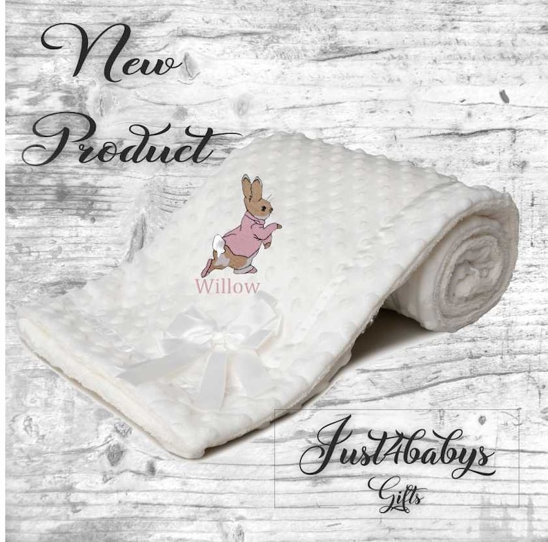 May include: White baby blanket with a white satin bow and a pink embroidered Peter Rabbit with the name "Willow" embroidered below. The blanket is rolled up and the text "Just4babys Gifts" is visible in the bottom right corner.