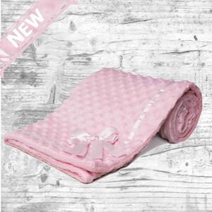 May include: A pink baby blanket with a white satin trim and a pink satin bow. The blanket is made of soft, plush fabric and has a textured surface.