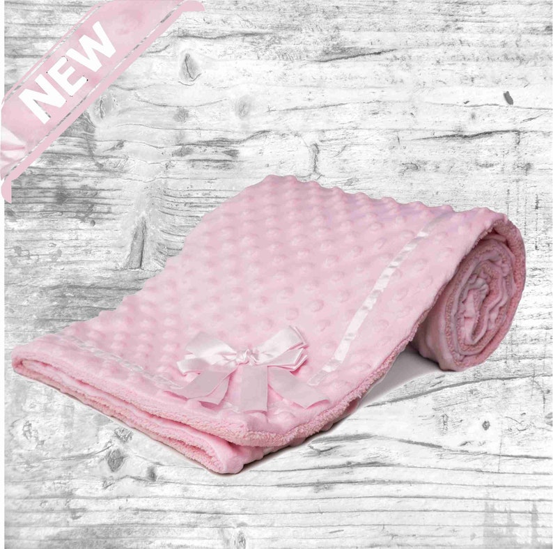 May include: A pink baby blanket with a white satin trim and a pink satin bow. The blanket is made of soft, plush fabric and has a textured surface.