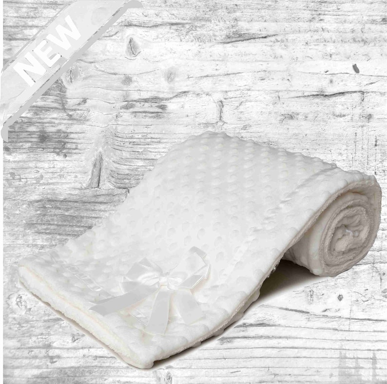 May include: White baby blanket with a satin bow. The blanket is made of soft, plush fabric and has a dotted pattern.