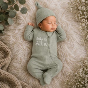 May include: A sage green ribbed long-sleeve footed sleeper with a matching knotted hat. The sleeper features a front zip fastening and white embroidered text that reads baby Albie with a small heart icon above the name on the chest area.