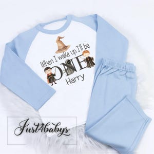 May include: A baby's two-piece pyjama set. The top is white with light blue sleeves and features a graphic with the text "When I wake up I'll be ONE Harry" and illustrations. The matching trousers are light blue.