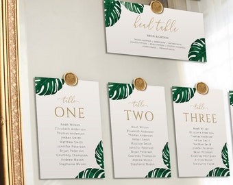 May include: A wedding seating chart with a tropical theme. The chart is displayed on a mirror with a gold frame. The cards are decorated with green monstera leaves and gold lettering. The cards are labeled with table numbers from one to eight. Each card lists the names of the guests assigned to that table.