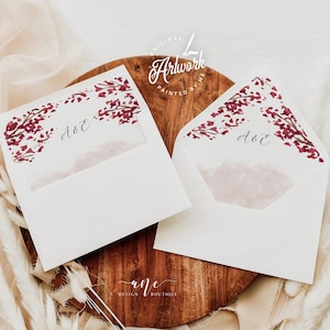 May include: Two white envelopes with a burgundy floral design and the initials "A & E" written in calligraphy. The envelopes are on a wooden surface.