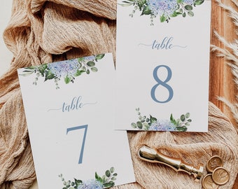 May include: Two watercolor table number cards with blue and green floral designs. The cards are on a wooden surface with a tan fabric and a gold wax seal. The text on the cards reads "table 7" and "table 8".