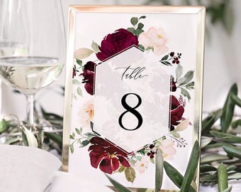 May include: A gold-framed table number card with a white background and burgundy and blush floral design. The card features a hexagonal frame with the text "Table 8" in black.