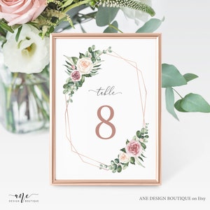 May include: A white card with a gold geometric frame and floral accents. The card says "table 8" in a rose gold font.