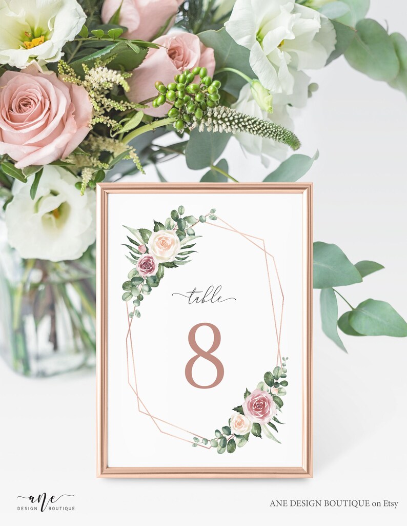 May include: A white card with a gold geometric frame and floral accents. The card says "table 8" in a rose gold font.