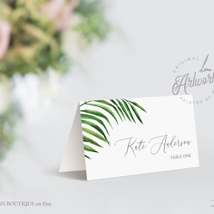 May include: White place card with a green palm leaf design. The text "Kete Anderson" and "Table One" is printed in black.