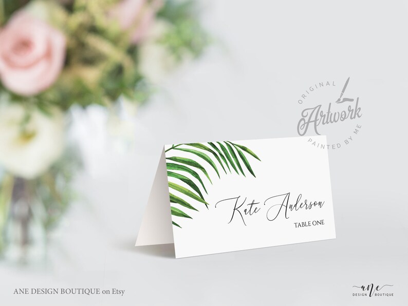 May include: White place card with a green palm leaf design. The text "Kete Anderson" and "Table One" is printed in black.