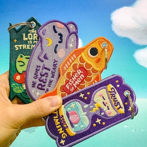 May include: Four embroidered patches with keychains. The patches are purple, green, orange, and light purple. The patches have various sayings on them, including "The Lord is my strength", "He gives rest to the weary", "Fisher of Men", and "God's Timing + Trust".