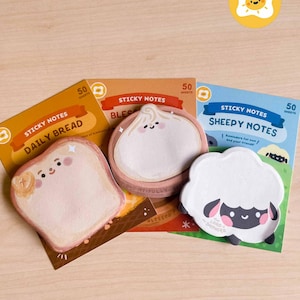 May include: A collection of cute sticky notes in various shapes and designs. The notes include a slice of bread, a dumpling, and a sheep, each with a cheerful face. Each pad contains 50 sheets. The notes are on a light brown wooden surface.