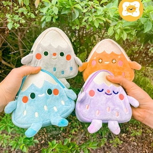 May include: Four plush mountain-shaped coin purses in gray, orange, blue, and purple. Each has a smiling face, arms, and legs. The purses have a zipper closure and are held against a background of green foliage.