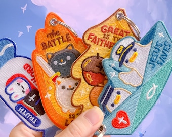 May include: Embroidered keychains in various shapes and colors. One keychain features a bear with the word "TRUTH", another has a cat with the text "the BATTLE", and a third has an acorn with the text "GREAT IS THE FAITH". The last keychain has a duck with the text "JESUS SAVES".