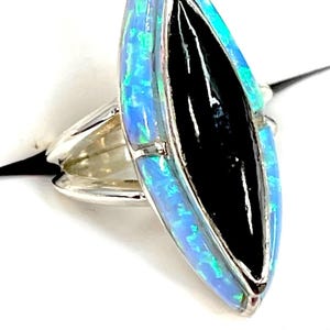 May include: A marquise-shaped ring featuring a central black stone inlay flanked by two bright blue iridescent opal segments. The ring is set in a polished silver-tone metal band that splits into two distinct prongs at the sides of the setting.