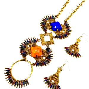 May include: A gold-tone necklace and earring set featuring a chain with a central square link, a blue square stone, an orange square stone, and a circular pendant. The pieces are decorated with fan-shaped accents containing small red, blue, and green stones.