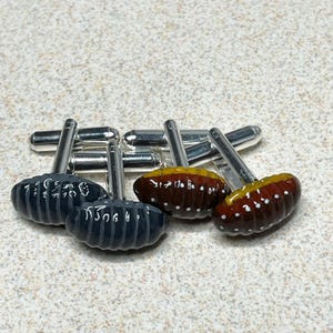 May include: A set of four cufflinks with silver-toned stems. Two pairs feature oval-shaped designs, one in dark gray with etched details, and the other in brown with yellow accents and white dots.