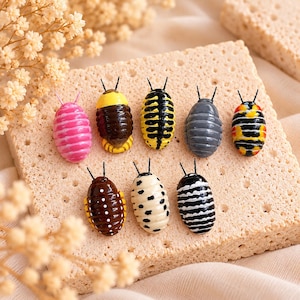 May include: A collection of eight small, oval-shaped figurines resembling pill bugs or woodlice, each featuring distinct patterns and colours including pink, yellow, black, grey, brown, and white, all with two thin antennae extending from the top.
