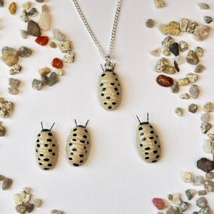 May include: A silver chain necklace with a pendant shaped like a beige insect with black spots. Three similar insect-shaped ornaments are also present, along with scattered pebbles and stones in various colors.