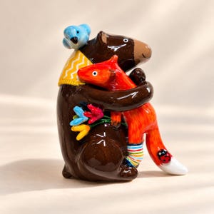 Op de afbeelding: Een glanzend keramieken beeldje van een donkerbruine beer met een gele sjaal met wit chevronpatroon, die een feloranje vos omhelst. De beer houdt een boeket kleurrijke bloemen vast en een blauwe vogel zit bovenop zijn hoofd.