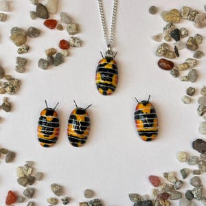 May include: A silver chain necklace with a pendant and three matching decorative figurines. The pendant and figurines are oval-shaped with a black, yellow, and orange striped design. Small black antennae are on each piece.