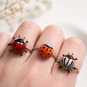 May include: Three rings with ladybug designs and silver bands. One ring has a red ladybug with black spots and antennae, another an orange ladybug with black spots, and the third a white and black striped ladybug.