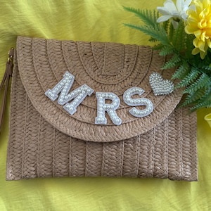 May include: A woven straw clutch in a light brown hue, featuring the word "MRS" spelled out in pearl-studded letters. A small pearl-studded heart is also on the clutch. The clutch has a wrist strap and a flap closure. The background is yellow.