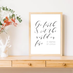 Puede incluir: Un cuadro enmarcado con la cita "Go forth & set the world on fire" en elegante escritura negra. El cuadro está en un marco de madera clara y se muestra sobre una superficie de madera. El texto incluye "St. Ignatius of Loyola".