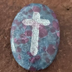 May include: A gray and purple oval-shaped stone with a white cross painted on it.