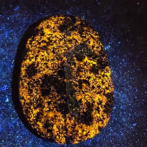 May include: A close-up of a dark, round, smooth stone with a bright yellow, speckled pattern. The stone is illuminated with a UV light, causing the yellow specks to glow.