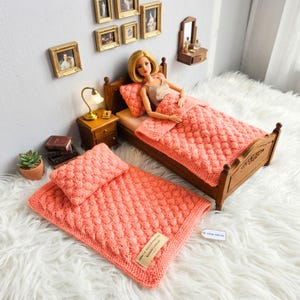 Op de afbeelding: Een miniatuur slaapkamer met een pop in een houten bed, bedekt met een koraalkleurige gebreide deken en kussen. Een tweede bijpassende deken en kussenset ligt op de vloer. De kamer omvat een nachtkastje, een lamp en ingelijste foto's aan de muur.